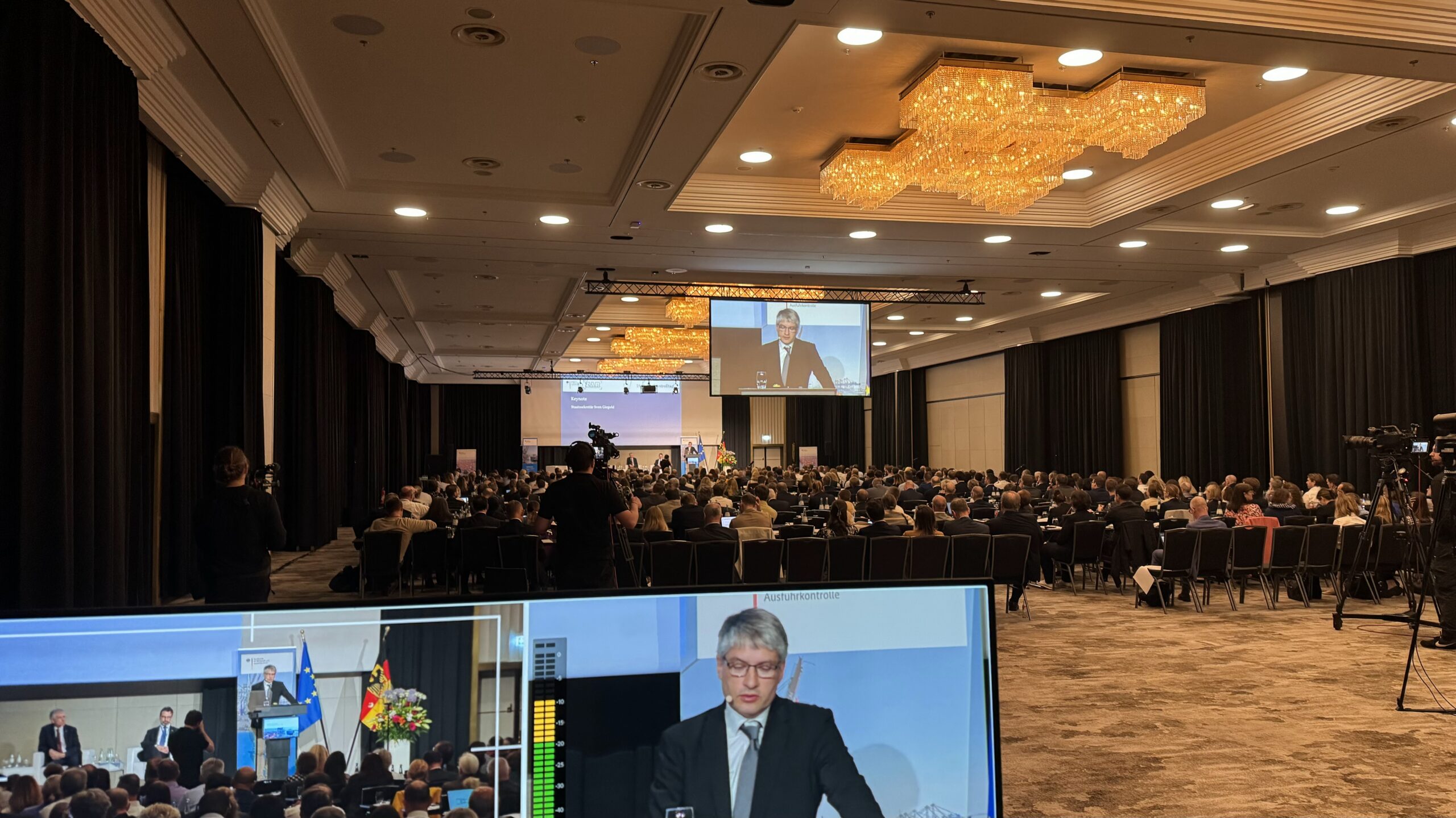 Großer Konferenzsaal mit Publikum, Redner auf Großbildschirm und professioneller Streaming-Technik mit Monitoren im Vordergrund.