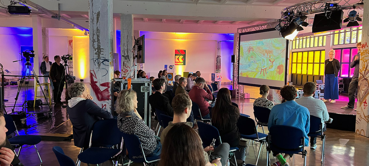 Videoproduktion kidsCon 2022: Kameras, Bühne mit Sprecher, großem Screen und Publikum in farbig beleuchteter Halle